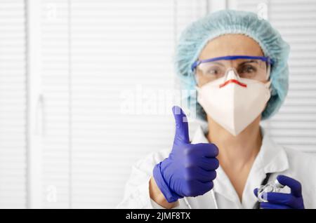 Ritratto di felice sorridente medico caucasico in maschera occhiali guanti e cappello mostra pollice su sfondo bianco finestra Foto Stock