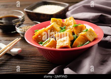 Croccante mescolare cubetti di tofu fritti con erba cipollina in piatto di argilla su tavolo da cucina in legno con tovagliolo e salsa di soia da parte Foto Stock