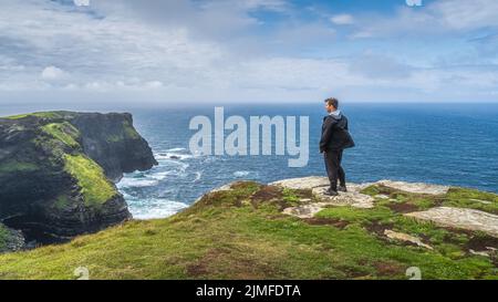 Uomo in piedi sulla cima delle iconiche scogliere di Moher e ammirando la vista spettacolare, l'Irlanda Foto Stock