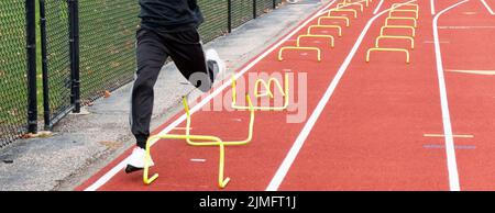 Un runner della scuola superiore viaggia sopra i mini hurdles gialli stabiliti su una pista per correre sopra durante il trapano del wicket alla pratica della pista. Foto Stock