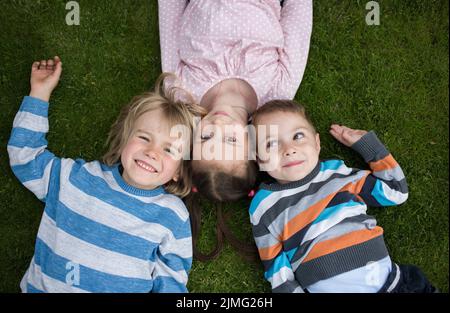 bambini felici, due ragazzi e una ragazza si trovano sull'erba con un senso di gioia e di unità. Buon umore, infanzia gioiosa spensierata, amicizia. Vista dall'alto Foto Stock
