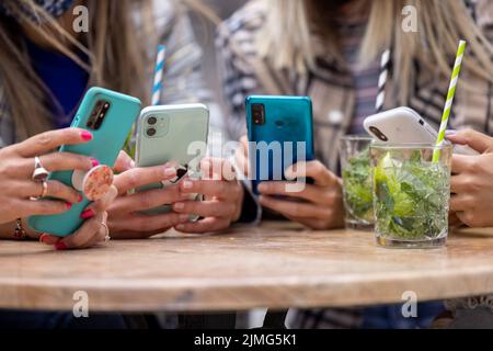 Primo piano delle mani di un gruppo multirazziale di quattro donne che utilizzano smartphone al tavolo in un bar della città con cocktail e dri Foto Stock