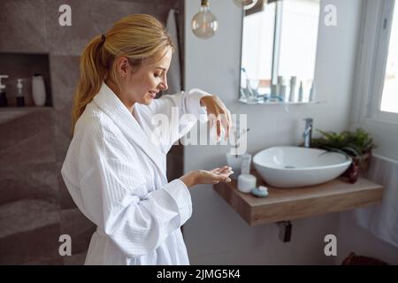 Giovane bella donna caucasica versando lozione corpo a portata di mano in bagno. Concetto anti-invecchiamento. Foto Stock