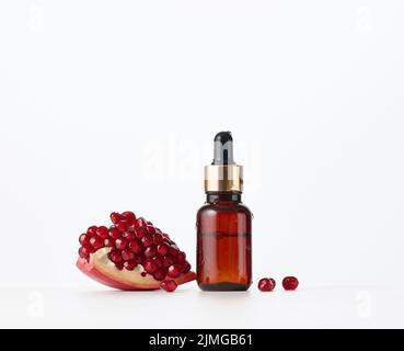 Bottiglia di vetro marrone con pipetta e semi di melograno rosso su fondo bianco. Modello per prodotti cosmetici liquidi, pubblicità Foto Stock