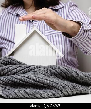 Mano femmina su una casa bianca in miniatura avvolta in una sciarpa grigia, concetto di isolamento edilizio Foto Stock