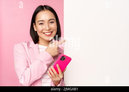 Immagine di una donna corporate asiatica sorridente in tuta, che tiene lo smartphone, indica la lavagna, mostra la carta o il logo delle informazioni sulla scritta Foto Stock