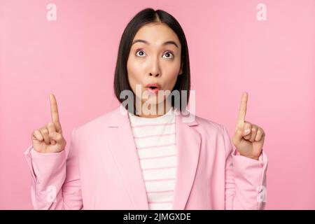 Entusiasta lavoratore aziendale, donna asiatica d'affari puntando le dita verso l'alto e sorridendo, mostrando pubblicità, logo, in piedi Foto Stock