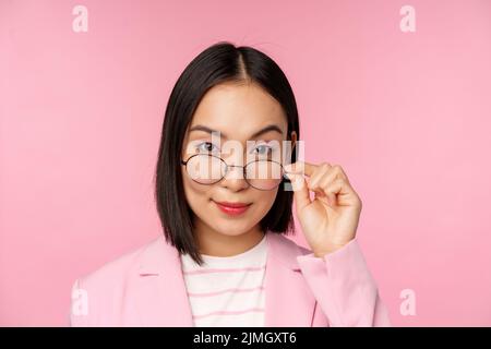 Ritratto di donna d'affari asiatica in occhiali, guardando incuriosito alla macchina fotografica e sorridente, professionista saleswoman fissando con interno Foto Stock