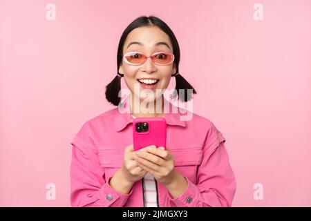 Ritratto di ragazza coreana in eleganti occhiali da sole che reggono il telefono cellulare, utilizzando l'app per smartphone, in piedi su sfondo rosa Foto Stock