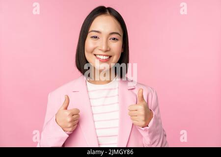 Donna d'affari professionale, donna asiatica d'affari che mostra pollici e sorridente, lode e complimento, in piedi in tuta su pi Foto Stock