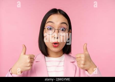 Primo piano ritratto di impressionato corporate woman, asiatica d'affari donna in occhiali, mostrando pollici in su, guardando stupito la macchina fotografica, rec Foto Stock