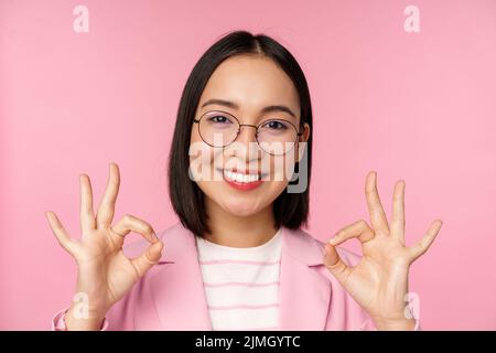 Primo piano ritratto di impressionato corporate woman, asiatica d'affari donna in occhiali, mostrando OK segno, guardando stupito la macchina fotografica, rec Foto Stock