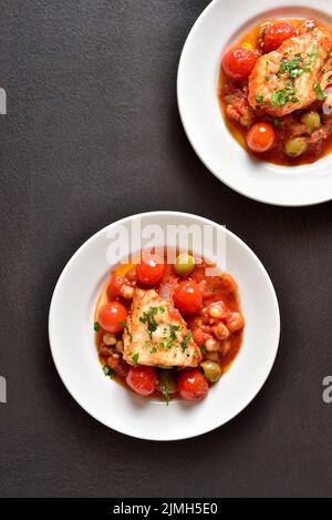 Stufato di merluzzo con ceci, pomodori ciliegini e olive sul piatto su sfondo di pietra scura. Vista dall'alto, piatto Foto Stock