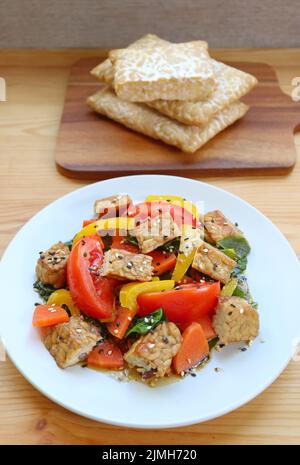 Insalata di verdure succulente e cubetti di tempeh arrosto con una pila di tempeh fresco sullo sfondo Foto Stock