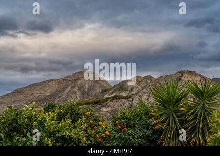 Un paesaggio montano desertico in Andalusia sotto cieli tempestosi con palme e frutteto arancione in primo piano Foto Stock