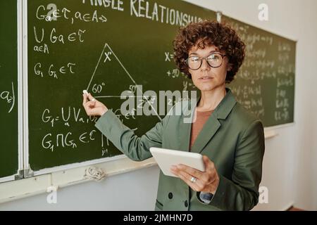 Insegnante sicuro di geometria che punta al triangolo sulla lavagna disegnato tra formule ed equazioni mentre spiega il nuovo soggetto Foto Stock