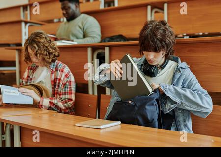 Ragazza bruna adolescente in casualwear mettendo il libro in zaino dopo lezione mentre si siede dalla scrivania in auditorium contro i suoi compagni di classe Foto Stock