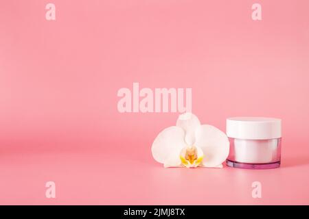 Vaso cosmetico in vetro con crema con fiore di orchidea isolato su sfondo rosa. Foto Stock