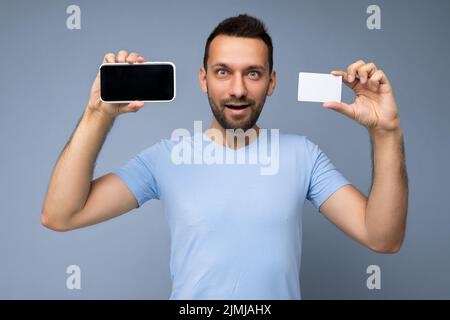 Foto di un bel giovane brunetto non rasato con barba che indossa una maglietta blu tutti i giorni isolata su sfondo blu che tiene e u Foto Stock