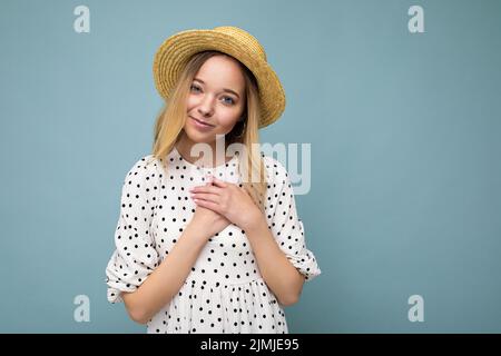 Foto di giovane positiva felice bella donna bionda con emozioni sincere indossando abito estivo e cappello di paglia isolato su blu Foto Stock