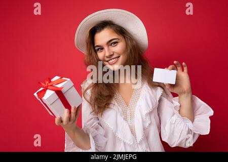 Giovane bella donna europea elegante brunetta indossando blusa bianca e cappello alla moda isolato su sfondo rosso con posit Foto Stock