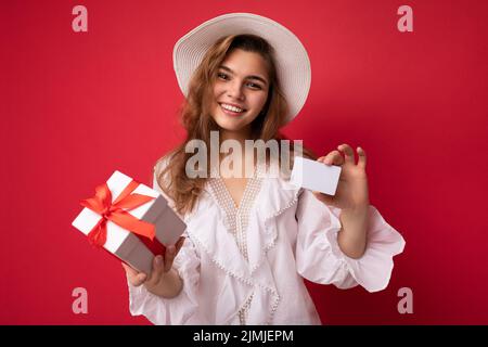 Ritratto di donna allegra positiva alla moda in formalwear con scatola regalo e carta di credito guardando la macchina fotografica isolato sul rosso Foto Stock