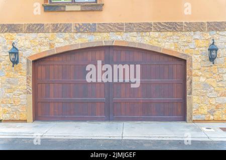 Doppio portoni in legno per garage con ingresso ad arco a Carlsbad, San Diego, California. Esterno garage con pareti laterali in legno di pietra e due lampade da parete Foto Stock