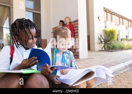 Sorridenti ragazzi della scuola elementare multirazziale che studiano mentre si siedono sul pavimento all'ingresso della scuola Foto Stock