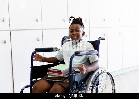 Ritratto di scolaro elementare afroamericano sorridente con libri seduti su sedia a rotelle Foto Stock