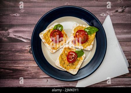 Tre cracker di riso con hummus di ceci, pomodori ciliegini e basilico fresco su un piatto su un tavolo rustico. Cibo sano. Foto Stock