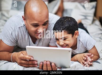 Questo è il programma che ti piace tanto: Un giovane allegro e suo figlio guardano i video su un tablet digitale mentre si aggrappano a un letto a casa Foto Stock