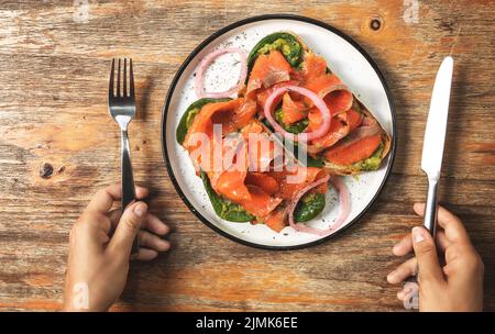 Mani femminili e toast deliziosi con salmone affumicato, avocado e foglie di basilico Foto Stock