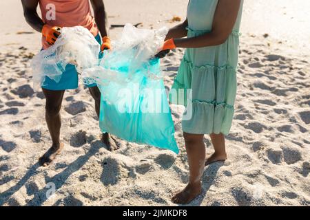 Sezione bassa di coppia afroamericana che raccoglie i rifiuti di plastica in sacchetto alla spiaggia il giorno di sole Foto Stock