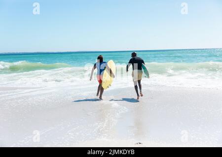Coppia di anziani afroamericani che trasportano tavole da surf che corrono verso onde in spiaggia con spazio copia Foto Stock