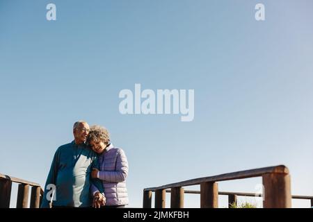 Amorevole coppia anziana sorridendo e tenendo le mani mentre si cammina lungo un ponte pedonale sul mare. Coppia anziana affettuosa che gode di una spiaggia rilassante Foto Stock