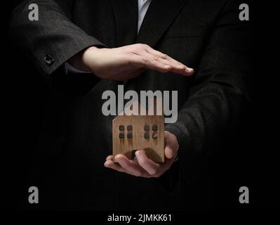 Uomo d'affari in tuta tiene un modello di legno di una casa su uno sfondo scuro. Concetto di vendita e acquisto immobiliare, affitto. Insuran Foto Stock