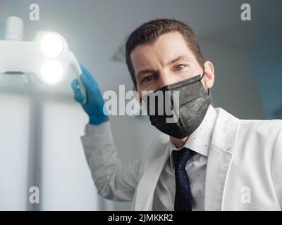 Ispezione presso il dentista dal punto di vista del paziente. Dottore che regola le luci operatorie e tiene lo specchio dentale in ufficio dentale Foto Stock