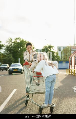 La madre felice e le sue figlie stanno avendo divertimento con un carrello di shopping su un parcheggio accanto ad un supermercato. Foto Stock