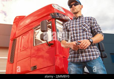 Caucasico Trucker nel suo 40s orgogliosamente in piedi di fronte alla cabina Rossa del suo veicolo Heavy Goods in attesa di Cargo per partire. Trasporti e logistica Foto Stock