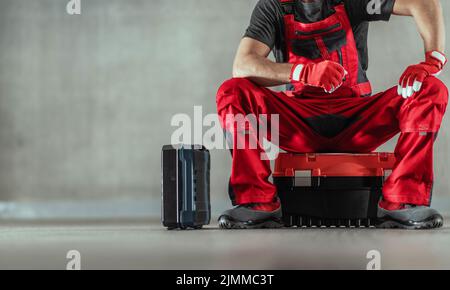 Primo piano di Handyman in Red Guanti da lavoro funzionali e protettivi seduti su una delle sue cassette degli attrezzi in attesa del cliente nella parte interna del viso Foto Stock