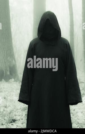 Morte in una felpa nera con cappuccio nella nebbia di nebbia di nebbia di foresta. Stile orrore paura spaventoso male Foto Stock