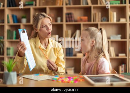 Cheerful il terapista femminile di discorso che cura i problemi e gli impedimenti della ragazza, apprendimento del bambino cute e le lettere pronuncianti Foto Stock