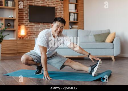 Felice asiatico uomo maturo formazione a casa, allungando le gambe in soggiorno interno, godendo di stile di vita sano Foto Stock