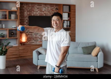 Sport domestici. Felice uomo asiatico di mezza età facendo esercizi di fitness con pesi, utilizzando manubri e sorridente Foto Stock