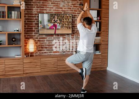 Vista posteriore di un uomo maturo che fa yoga mentre guarda il video di allenamento in TV, in piedi in posizione albero con le braccia sollevate Foto Stock