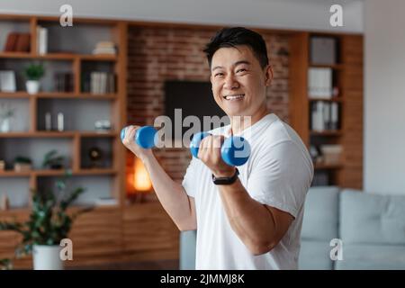 L'uomo maturo coreano allegro che fa esercizi di forza per le braccia, sollevando manubri in salotto, sorridendo alla macchina fotografica Foto Stock