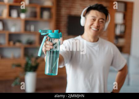Sorridente uomo asiatico attivo di mezza età in abbigliamento sportivo indossando cuffie e tenendo una bottiglia d'acqua, fuoco selettivo Foto Stock