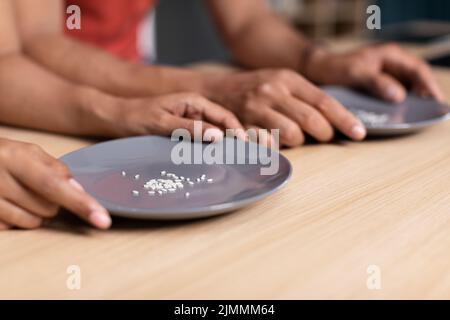 Mani di giovane famiglia nera tenere piatti con alcuni grani di riso a tavola in cucina, tagliato Foto Stock