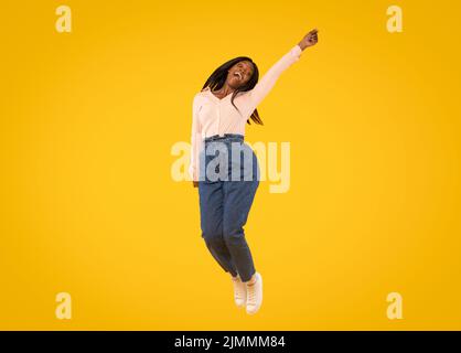 Gioiosa signora afroamericana che salta in aria e guarda la macchina fotografica, donna che si diverte su sfondo giallo studio Foto Stock