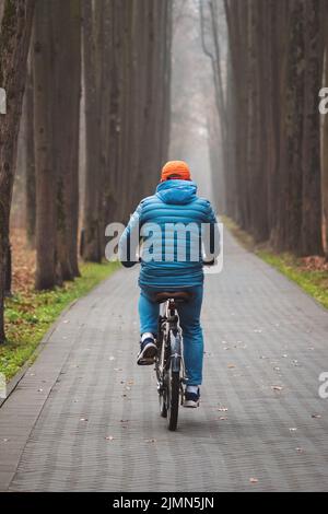 L'uomo va in bicicletta il fine settimana autunnale in un parco di nebbia opaco, vista posteriore Foto Stock